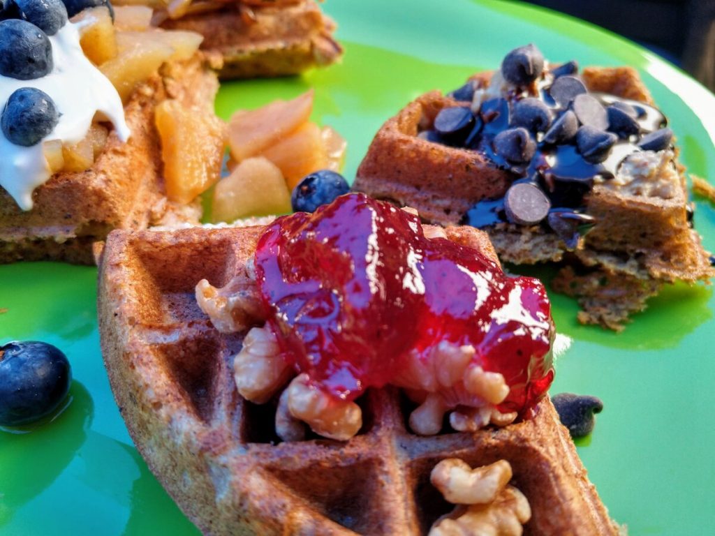 vegan waffle topped with walnuts, fruit jam, chocolate chips, blueberries, baked apples, and chocolate syrup
